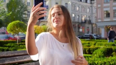 4k closeup video of beautiful young woman with long hair sitting on bench in city park and making selfie on smartphone camera