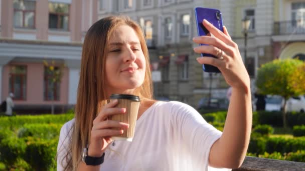 Портрет красивої молодої жінки з довгим волоссям, що робить селфі на камеру смартфона з кавою, щоб піти в паперову чашку. Модний блогер створює імідж для соціальних медіа — стокове відео