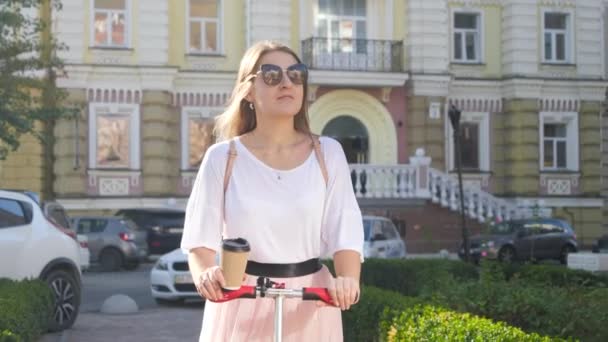 4k video de hermosa mujer sonriente montando scooter en la calle de la ciudad y beber café para ir en taza de papel — Vídeo de stock