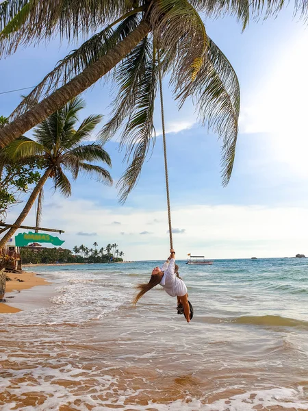 Красивая молодая женщина, качающаяся на веревке, привязанной к кокосовой пальме на берегу океана — стоковое фото