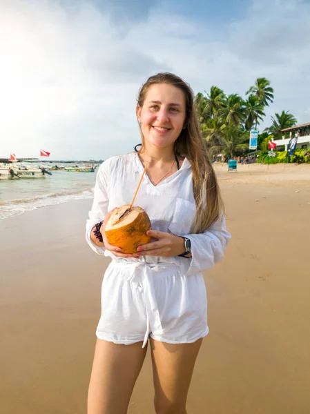 Портрет красивой улыбающейся молодой женщины, позирующей с кокосом на берегу океана на закате — стоковое фото