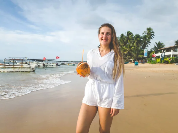 Портрет улыбающейся молодой женщины, держащей кокос с соломой на берегу океана — стоковое фото