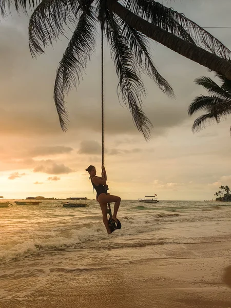 Krásná mladá žena v plavkách houpající se na laně přes oceánské vlny při západu slunce — Stock fotografie