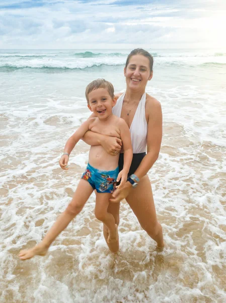 Retrato de feliz madre riendo sacando a su hijo sonriente del agua del océano —  Fotos de Stock