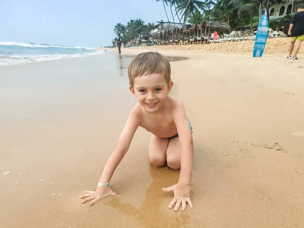 Portret uśmiechniętego chłopca siedzącego na mokrym piasku na plaży oceanicznej — Zdjęcie stockowe