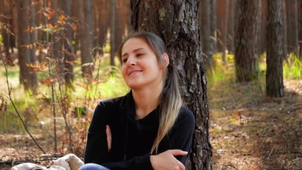 4k видео, где красивая улыбающаяся женщина отдыхает, сидя на земле в лесу или парке и опираясь на елку — стоковое видео