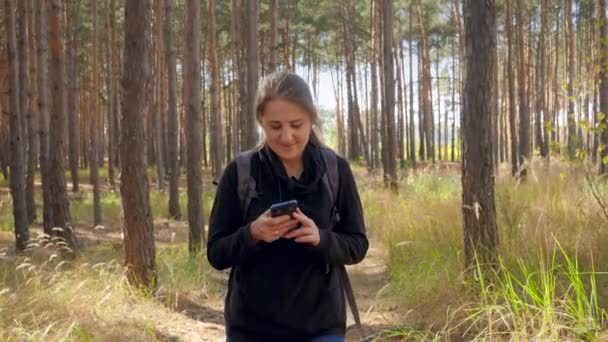 Vídeo 4k de bela jovem sorridente caminhando na floresta e digitando mensagem no smartphone ou olhando no mapa GPS enquanto caminha no caminho entre árvores altas — Vídeo de Stock