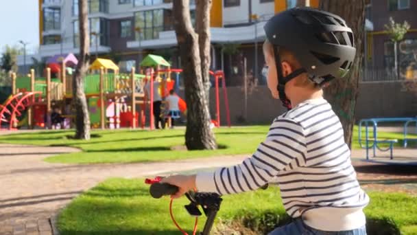Closeup 4k video of little boy wearing protective helmet riding on bicycle in park next to big children playground — ストック動画