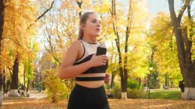 Closeup 4k video of beautiful young woman holding smartphone and listening to music while running and doing fitness exercise at autumn park