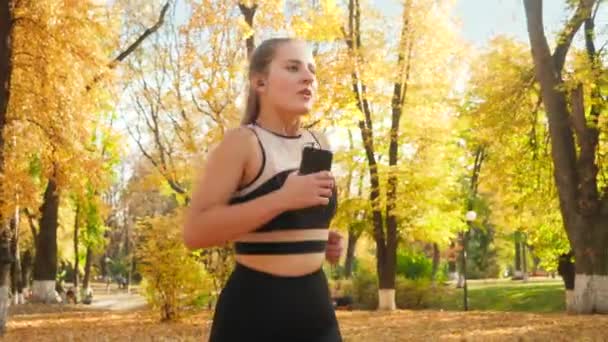 Closeup 4k video of beautiful young woman holding smartphone and listening to music while running and doing fitness exercise at autumn park — ストック動画