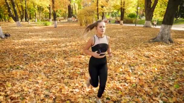 4k повільне відео руху з високої точки красивої молодої жінки в легінсах біжить на осінньому парку і слухає музику з навушниками — стокове відео