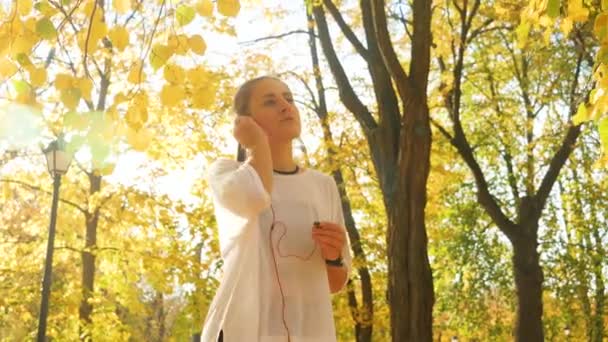 Vidéo au ralenti 4k de belle fille souriante mettant des erphones dans l'oreille et faisant du jogging dans un beau parc d'automne 