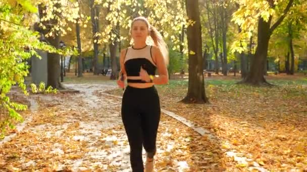 4k video of beautiful young woman in sexy sports unifor running on the pathway at autumn park under big trees — ストック動画
