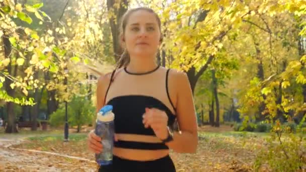 Portrait of beautiful smiling young woman running at park and drinking water from bottle — Stock video