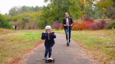 4k video of 4 years old smiling boy riding on scooter with his mother at autumn park