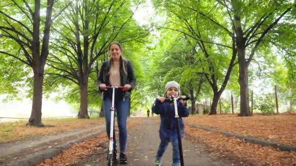 4k metraje de niño sonriente feliz con madre joven montando en scooters en hermoso callejón con árboles altos en el parque — Vídeo de stock