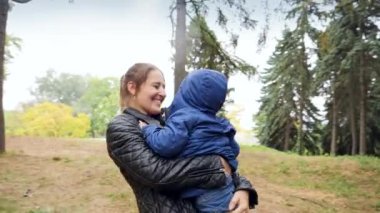 Closeup 4k video of happy smiling young mother hugging and embracing her little son under high trees at autumn park