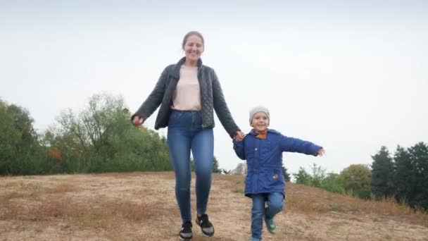 4k video de feliz riendo joven madre sosteniendo a su pequeño hijo a mano y corriendo en el prado en el parque de otoño — Vídeo de stock