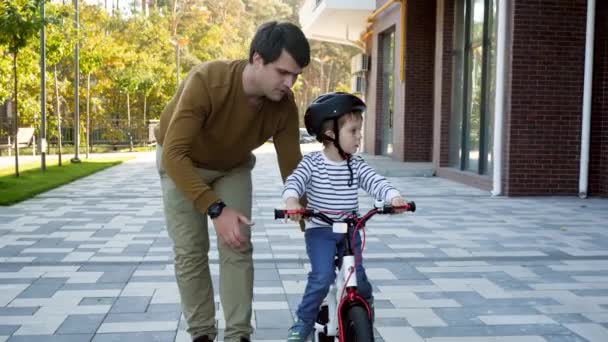 Imágenes de 4k del niño feliz usando casco protector aprendiendo a montar en bicicleta con su padre joven en la calle de la ciudad — Vídeos de Stock