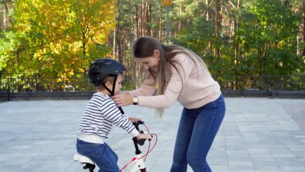 4k footage of young caring mother putting on and fastening protective bicycle helmet on her little son at park — Αρχείο Βίντεο