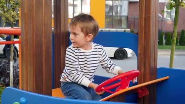 4k video of little boy pretending to be a driver and rotating steering wheel while playing on playground at park — ストック動画