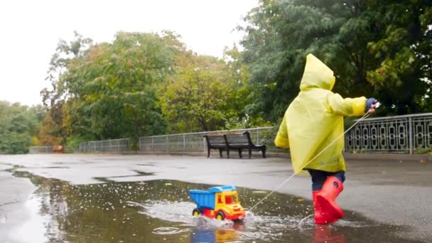 Відео з повільним рухом щасливого маленького хлопчика в гумових чоботях біжить і грає у великій калюжі з іграшковою вантажівкою під час дощу на осінньому парку — стокове відео