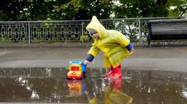 Slow motion video of little toddler boy in rubber boots and raincoat plying with toy truck in big puddle after raining at park
