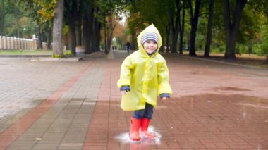 Slow motion video of happy laughing little boy in red wellington boots jumping in big puddle and spalshing water while raining at autumn park