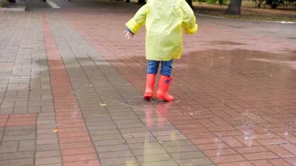 Slow motion video of little boy in red rubber boots and yellow raincoat jumping in big puddle after rain — 图库视频影像
