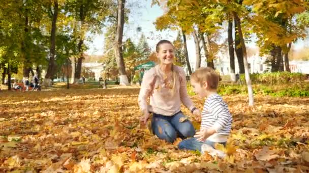 4k video of cheerful family with little boy sitting on grass at park and throwing yellow and red tree leaves — Stockvideo