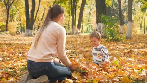 Vidéo 4k de joyeux garçon heureux avec sa mère assise au parc d'automne et vomissant des feuilles d'or 