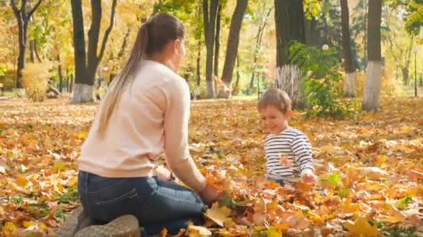 4k видео веселого счастливого мальчика с матерью, сидящей в осеннем парке и рвущей золотые листья — стоковое видео