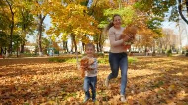 4k footage of happy msiling mother with 4 years old little son running in park and throwing autumn leaves in camera