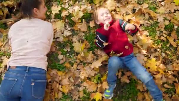 Vídeo 4k de mãe sorrindo e rindo com o filho pequeno rolando na grama coberta com folhas de árvore caídas amarelas no parque de outono — Vídeo de Stock