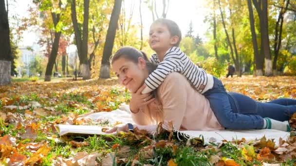 4k video of little boy climbing on mothers back and kissing her while she was lying on grass and yeallow leaves at autumn park — Stockvideo