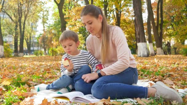 4k footage of young mother reading book and ating apple to her little son while having picnic in fall park — Wideo stockowe