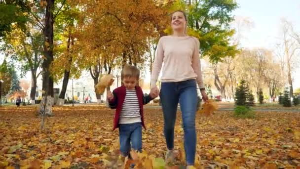 4k видео счастливой улыбающейся матери с маленьким сыном, гуляющим по траве, покрытой желтыми листьями в осеннем парке — стоковое видео