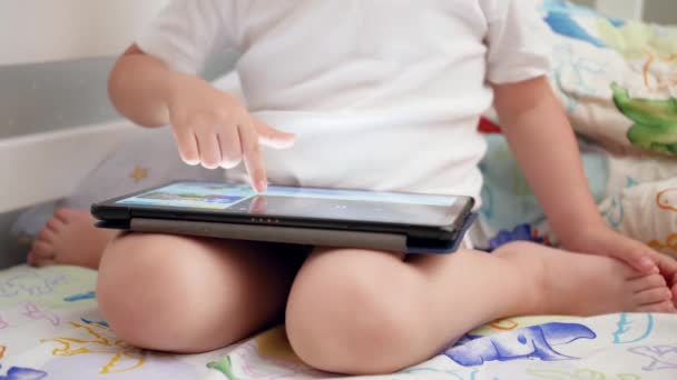 4k primer plano de vídeo de niño pequeño sentado en la cama y presionando la pantalla táctil de la tableta digital con el dedo — Vídeos de Stock