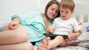 4k video of little toddler boy with young mother wearing pajamas lying in bed and playing games on tablet computer before going to sleep