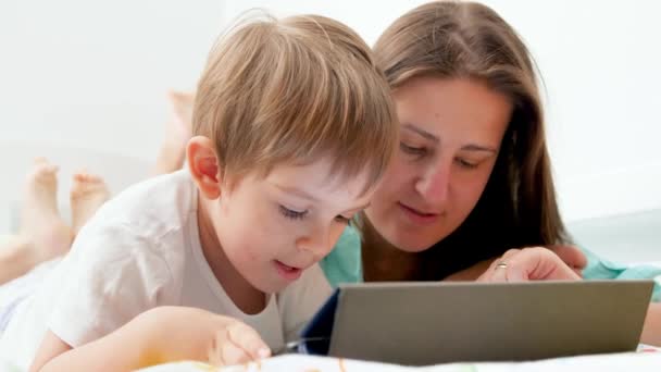 4k primer plano de vídeo de la madre joven acostada con su hijo pequeño y enseñándole a usar tableta y jugar juegos educativos — Vídeos de Stock