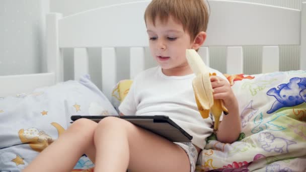 4k video of cute little boy lying in bed and eating banana while using digital tablet computer — Stockvideo