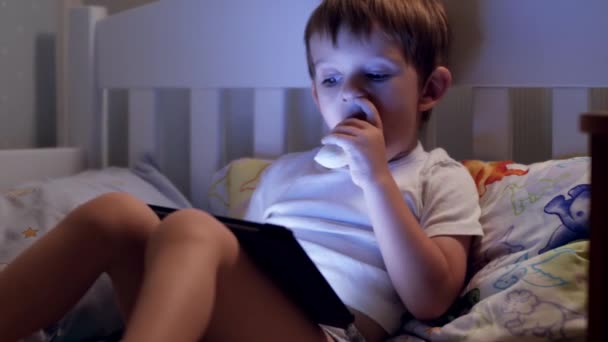 4K closeup video of little toddler boy watching cartoons on digital tablet computer and eating banana before going to sleep at night — Stok video