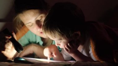 Kapalı 4K video, küçük çocuk gece annesiyle yatakta uzanıyor ve meşaleyle kitap okuyor.