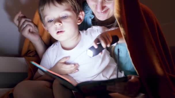 4k vídeo de feliz sorrindo mãe com o pequeno filho sentado sob cobertor à noite e lendo livro com lanterna — Vídeo de Stock