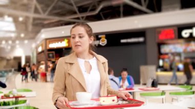 Elinde yemek tepsisi olan genç bir kadının alışveriş merkezinde yemek yemek ve oturmak için yer aradığı 4K video.