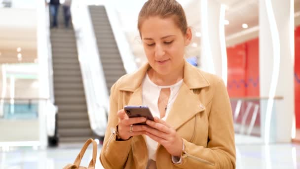 4k кадри красивої усміхненої жінки, що сидить на лавці поруч з ескалатором в торговому центрі і вводить повідомлення в соціальних мережах на смартфон — стокове відео