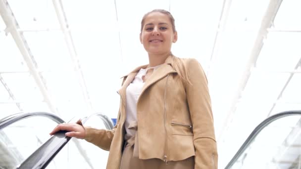 Vidéo 4k de belle jeune femme souriante descendant sur l'escalator dans le centre commercial et regardant à la caméra — Video