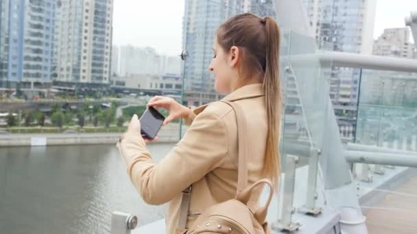 4k videó fiatal nő áll a tetőn, hogy a kép a város városkép és kaparók és kiküldetés a közösségi média segítségével okostelefon — Stock videók