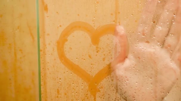 Closeup 4k video of young naked woman wiping heart shape drawn on the misted glass door at shower — Stockvideo