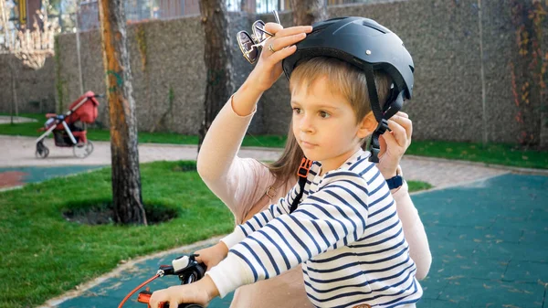 Foto de primer plano de la joven madre con casco protector en su hijo antes de montar en bicicleta — Foto de Stock
